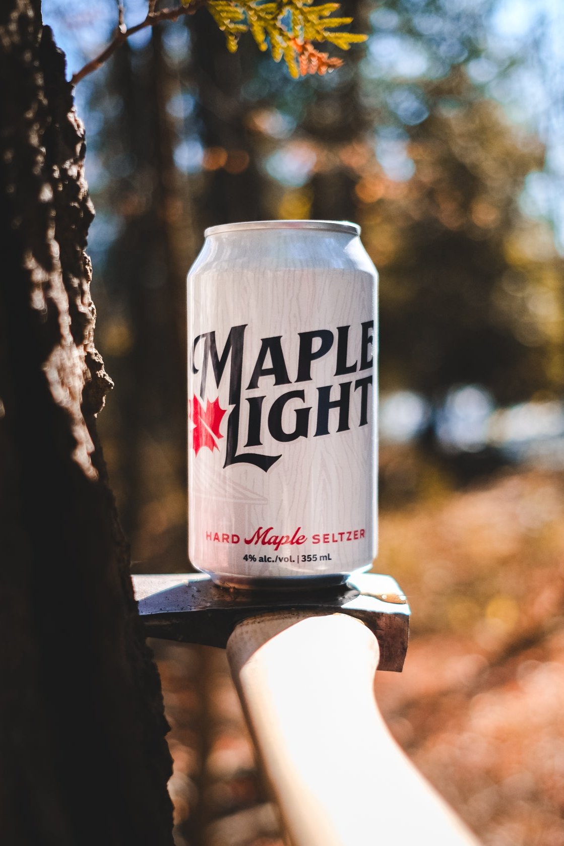 A can of Maple Light sitting on the head of an axe that's been wedged into the side of a tree.
