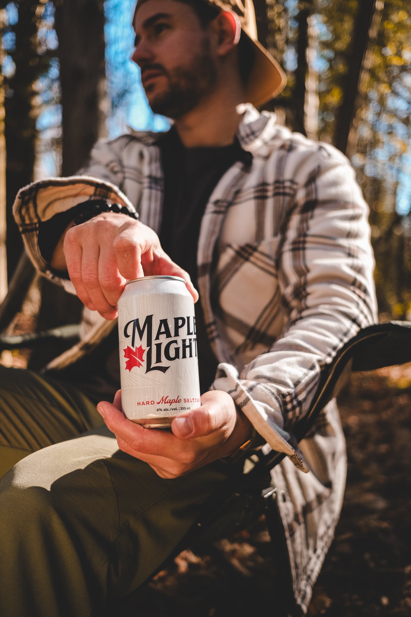 A man sitting in a camping chair in the forest cracking open a can of Maple Light.